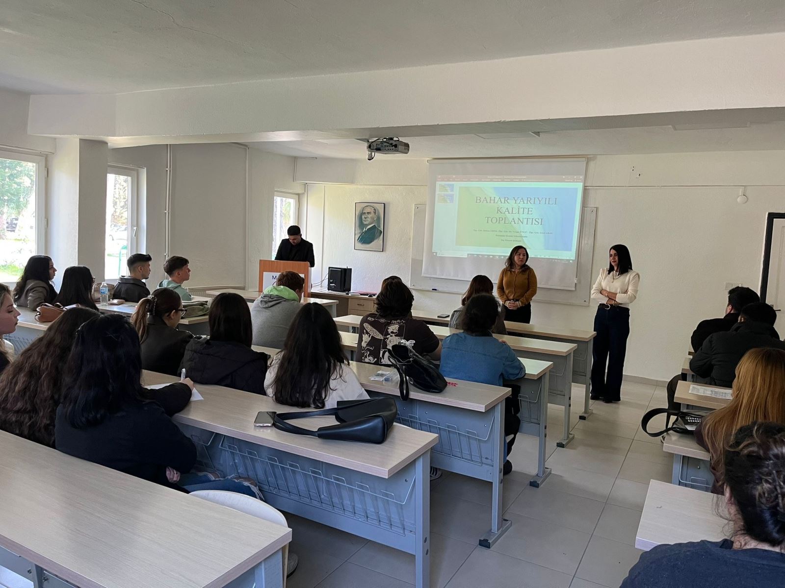 Bir üniversite sınıfında öğrencilerin katıldığı bir kalite toplantısı gerçekleştirilmektedir. Öğretim elemanları sınıfın önünde durarak öğrencilerle konuşmaktadır. Arkalarında projektör ekranında “Bahar Yarıyılı Kalite Toplantısı” başlıklı sunum yer almaktadır. Öğrenciler sıralarda oturmuş şekilde toplantıyı dikkatle takip etmektedir.